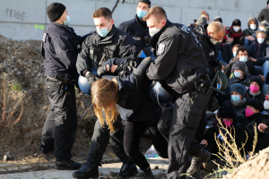 Gruppe maskierter Menschen in Mützen, einige mit Waffen, umringt eine Frau, die von der Polizei eskortiert wird, in einer städtischen Umgebung mit Pflanzen, Steinen und einer Wand mit Protesttext.