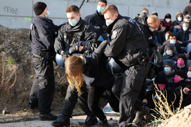 Gruppe maskierter Menschen in Mützen, einige mit Waffen, umringt eine Frau, die von der Polizei eskortiert wird, in einer städtischen Umgebung mit Pflanzen, Steinen und einer Wand mit Protesttext.