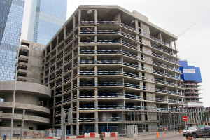Großes Bauwerk im Bau in einer Stadt mit Hochhäusern, Fahrzeugen, Fußgängern, Verkehrsinfrastruktur und Kränen vor einem klaren Himmel.