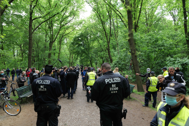 Polizisten in Uniform vor einer Menschenmenge mit Fahrrädern und einer Bank im Vordergrund und Bäumen und Himmel im Hintergrund.