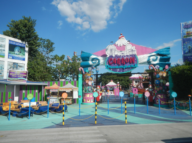 Karussell am Parkeingang mit Straße, Absperrung, Buden, Schildern, Bäumen und bewölktem Himmel im Hintergrund.
