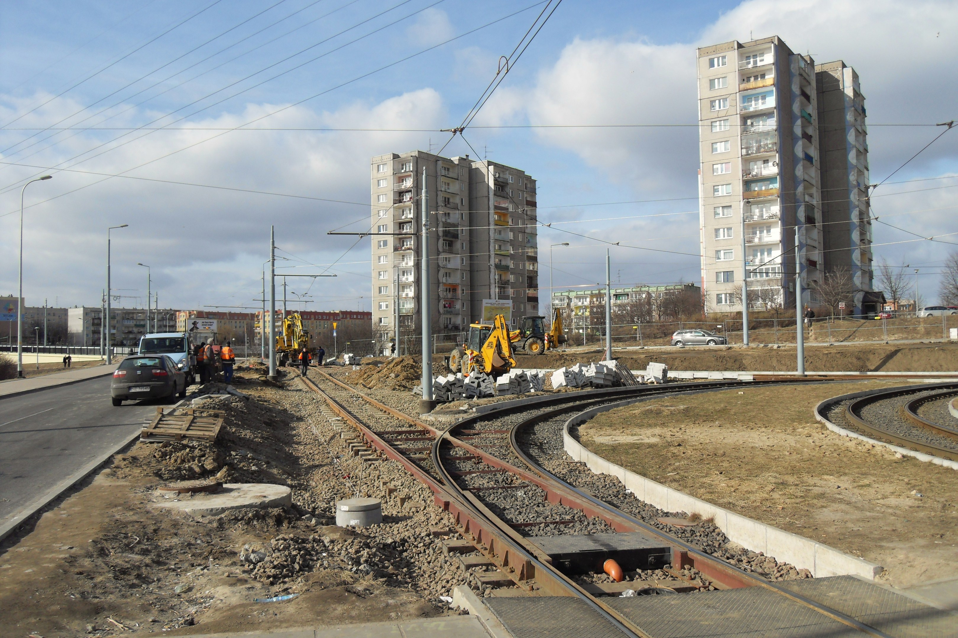 Baustelle mit einer Bahnschiene, Geb├Ąuden, Stra├čeninfrastruktur, Fahrzeugen, Passanten, Schutt, B├Ąumen und bew├Âlktem Himmel.