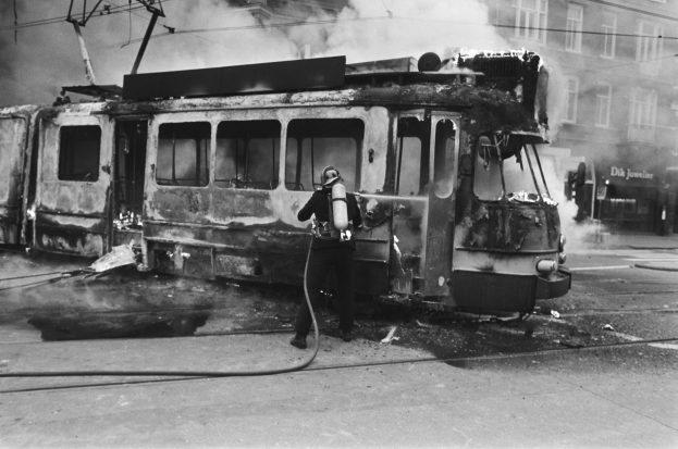 Schwarz-weißes Bild eines Mannes in einem Helm, der eine Pfeife h├Ąlt und neben einer ausgebrannten Stra├čenbahn mit aufsteigendem Rauch steht, auf einer Stra├če mit einem Geb├Ąude und Dr├Ąhten im Hintergrund.