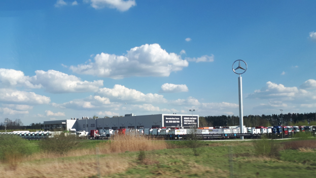 Eine Mercedes-Benz-Fabrik in Deutschland mit Fahrzeugen auf der Straße, Grünflächen, Gebäuden und einem Logo-Pfahl unter einem bewölkten Himmel.