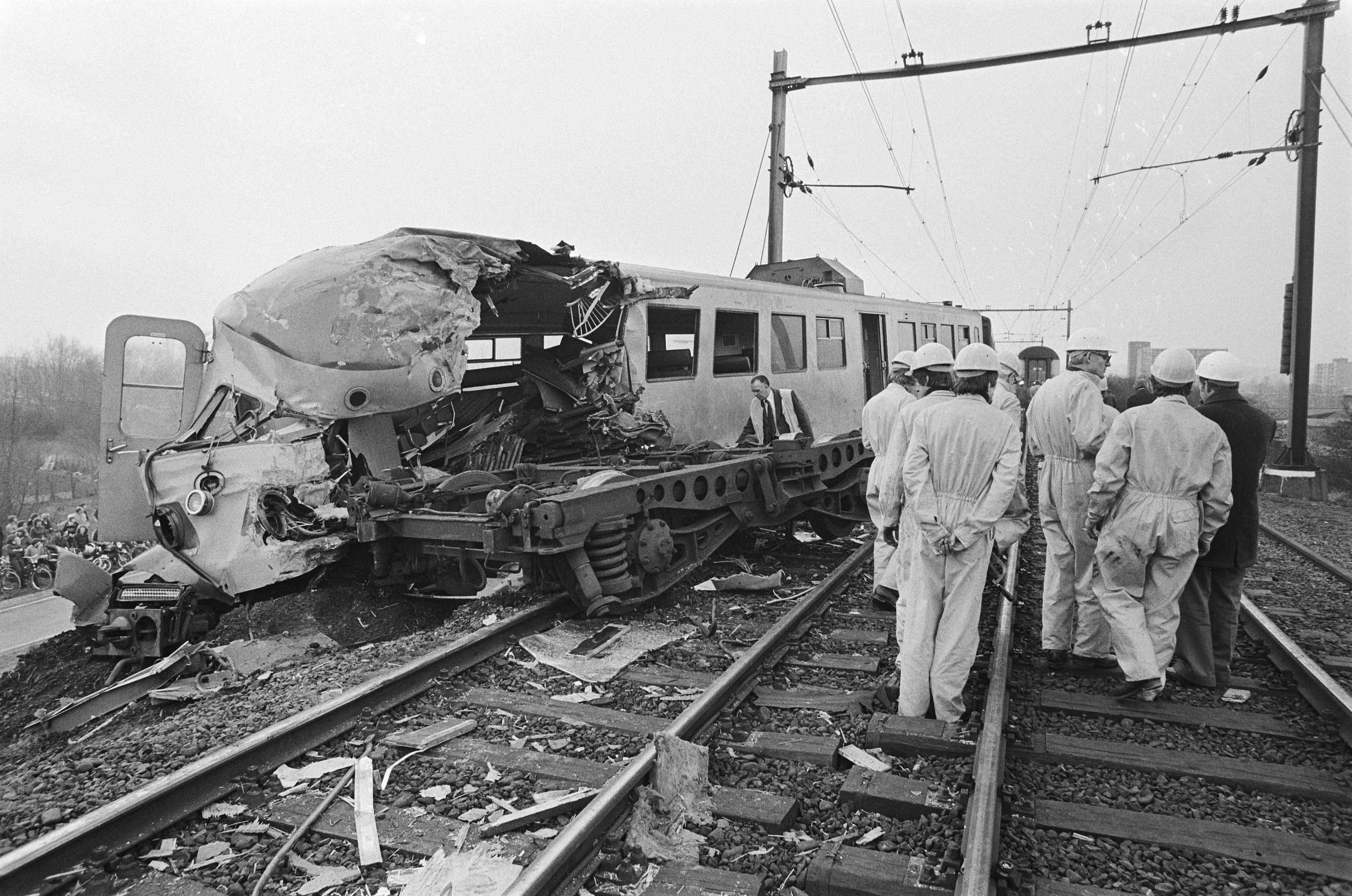 Schwarz-weißes Bild eines Zugunfalls auf den Schienen mit Einsatzkräften in Helmen und einem Hintergrund aus Strommasten, Bäumen, Gebäuden und einem klaren Himmel.