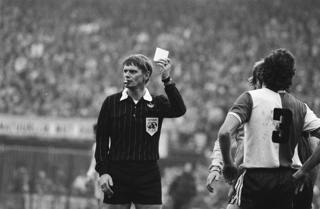 Schwarßt-weßts-Bild eines Schiedsrichters mit einer roten Karte vor einer Menge von Fußballspielern mit einem Zaun im Hintergrund.