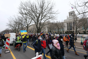 Eine große Gruppe von Menschen marschiert auf einer Straße in Washington, D.C. und hält Schilder und Banner, einige fahren Fahrräder, unter einem klaren blauen Himmel.