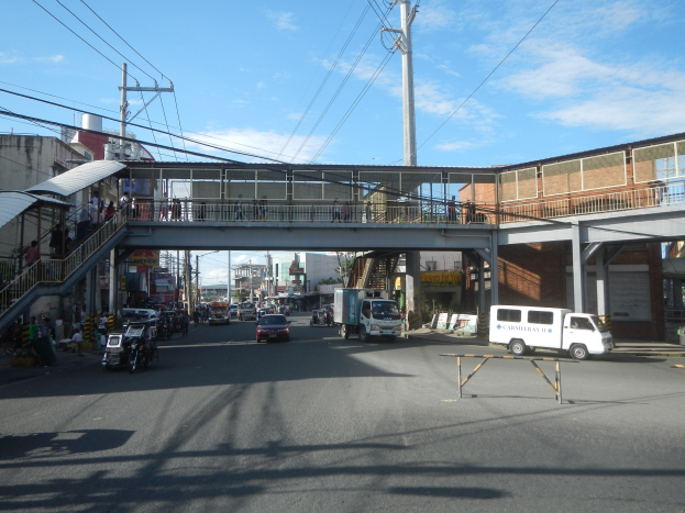 Stadtstraße mit Fahrzeugen, eine Fußgängerbrücke mit Menschen, Strommasten, Gebäude und ein bewölkter Himmel.