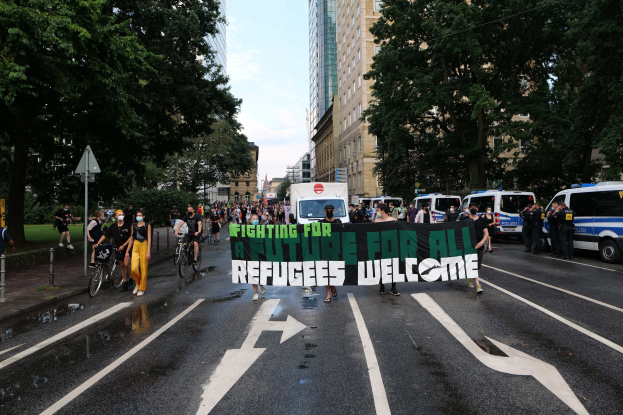 Eine Gruppe von Menschen, die eine Straße entlanggehen und ein Banner mit der Aufschrift "Flüchtlinge Willkommen" tragen, mit parkenden Fahrzeugen und Fahrradfahrern daneben, Bäumen, die die Straße säumen, und Gebäuden im Hintergrund.