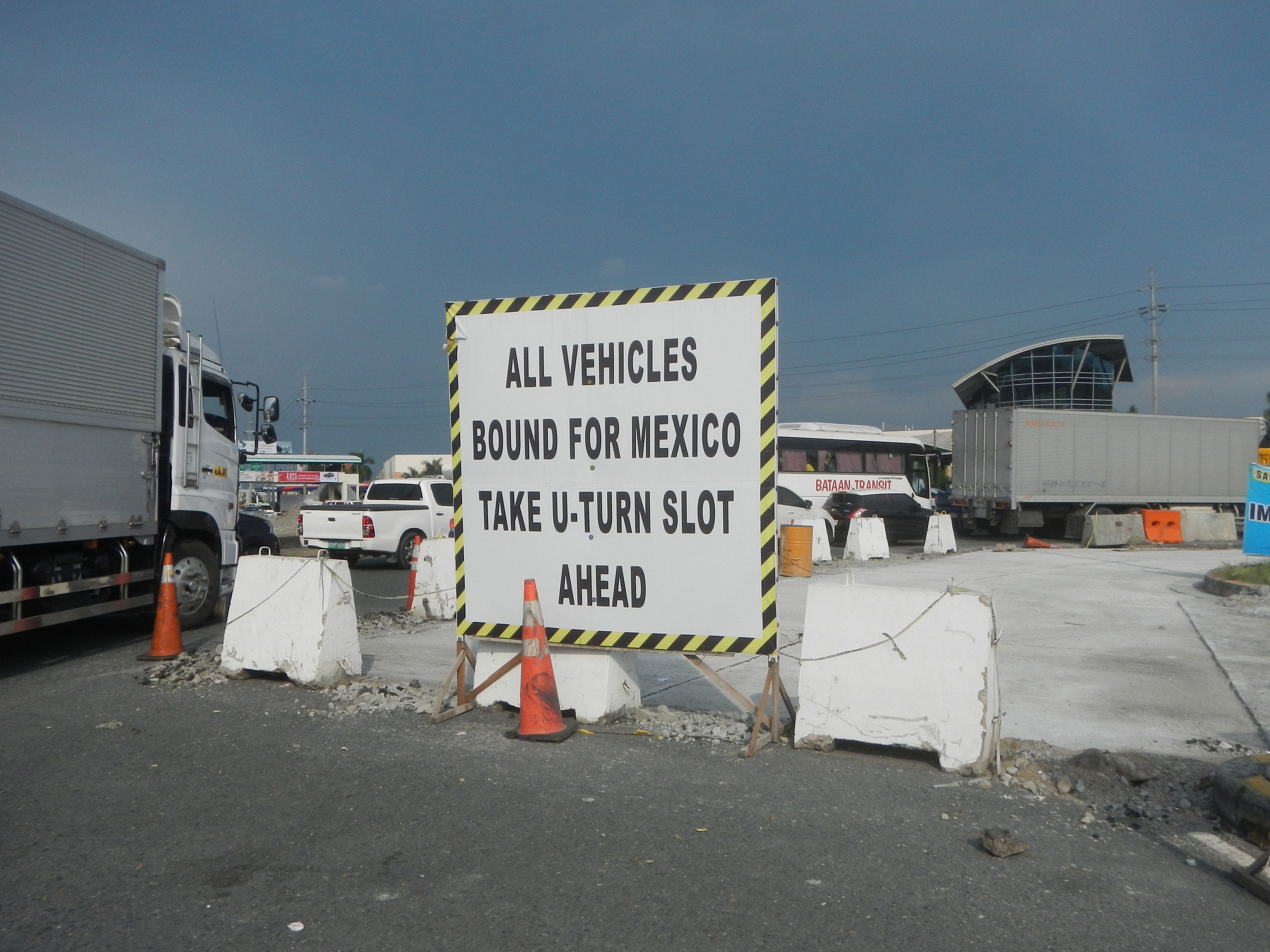 Ein Lastwagen auf einem Parkplatz neben einem Schild mit der Aufschrift "Alle Fahrzeuge nach Mexiko" geparkt, mit verstreuten Verkehrskegeln, Brettern und anderen Gegenständen und im Hintergrund Gebäude, Bäume, Pfosten und einen klaren blauen Himmel.