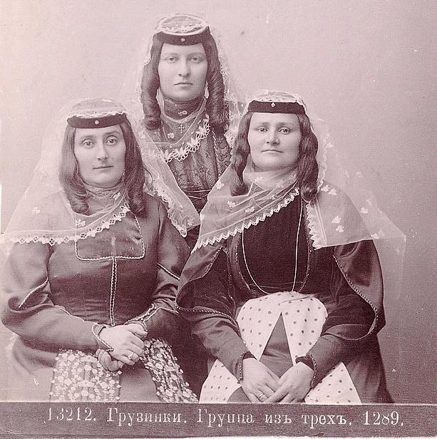 Drei Frauen in traditionellen russischen Hochzeitskleidern mit aufwendiger Stickerei und Perlenposen sich vor einer weißen Wand in einem Schwarz-Weiß-Foto.