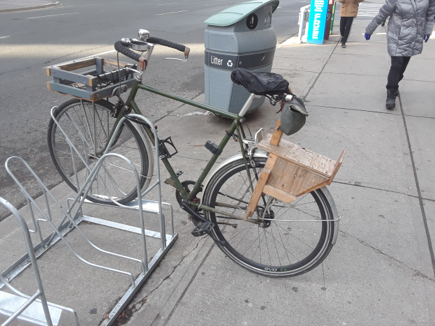 Ein grünes und braunes Fahrrad steht auf der Straße neben einem Müllcontainer, mit einigen Gegenständen daran befestigt, während Fahrzeuge auf der Straße fahren und Menschen auf dem Gehweg im Hintergrund zu sehen sind.