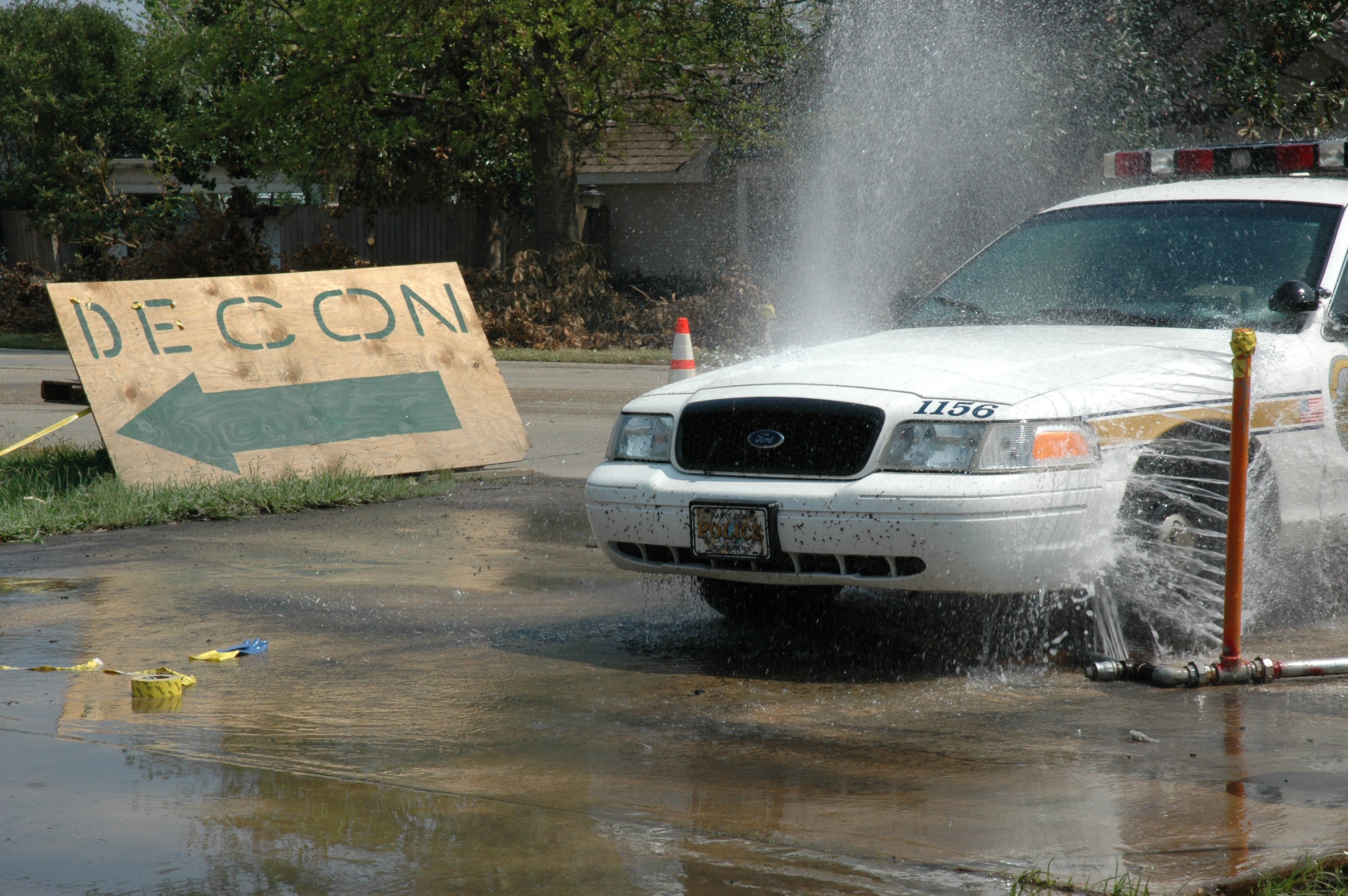 Polizeiauto fährt durch eine Pfütze mit verstreuter Trümmer, einschließlich einer Platte, eines Verkehrskegels und anderer Gegenstände; Bäume, Gebäude und ein klarer blauer Himmel im Hintergrund.