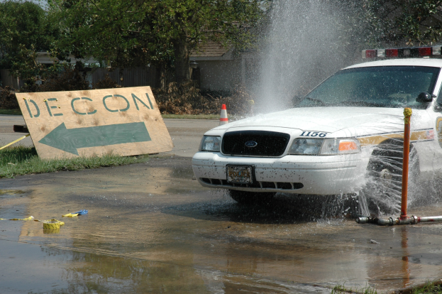 Polizeiauto fährt durch eine Pfütze mit verstreuter Trümmer, einschließlich einer Platte, eines Verkehrskegels und anderer Gegenstände; Bäume, Gebäude und ein klarer blauer Himmel im Hintergrund.
