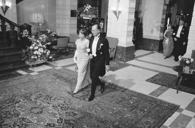 Ein Schwarz-Weiß-Bild einer Braut und eines Bräutigams, die durch eine Hotellobby gehen, umgeben von Menschen, Möbeln und Dekoration.