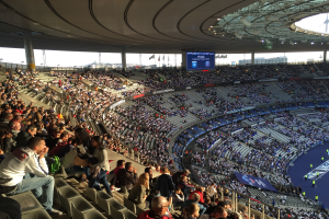 Große Zuschauermenge in einem Stadion bei einem Fußballspiel mit einer Bühne, Fahnen und einem Bildschirm im Hintergrund, identifiziert als Allianz Arena in München, Deutschland.