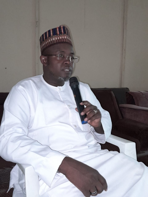 Ein Mann in einem weißen Kleid und einer Mütze sitzt in einem Stuhl und spricht in ein Mikrofon, mit zwei Sofas und einer Wand dahinter, die Wand hat die Wörter "Ich bin kein Lehrer, ich bin ein Lehrer" darauf geschrieben.