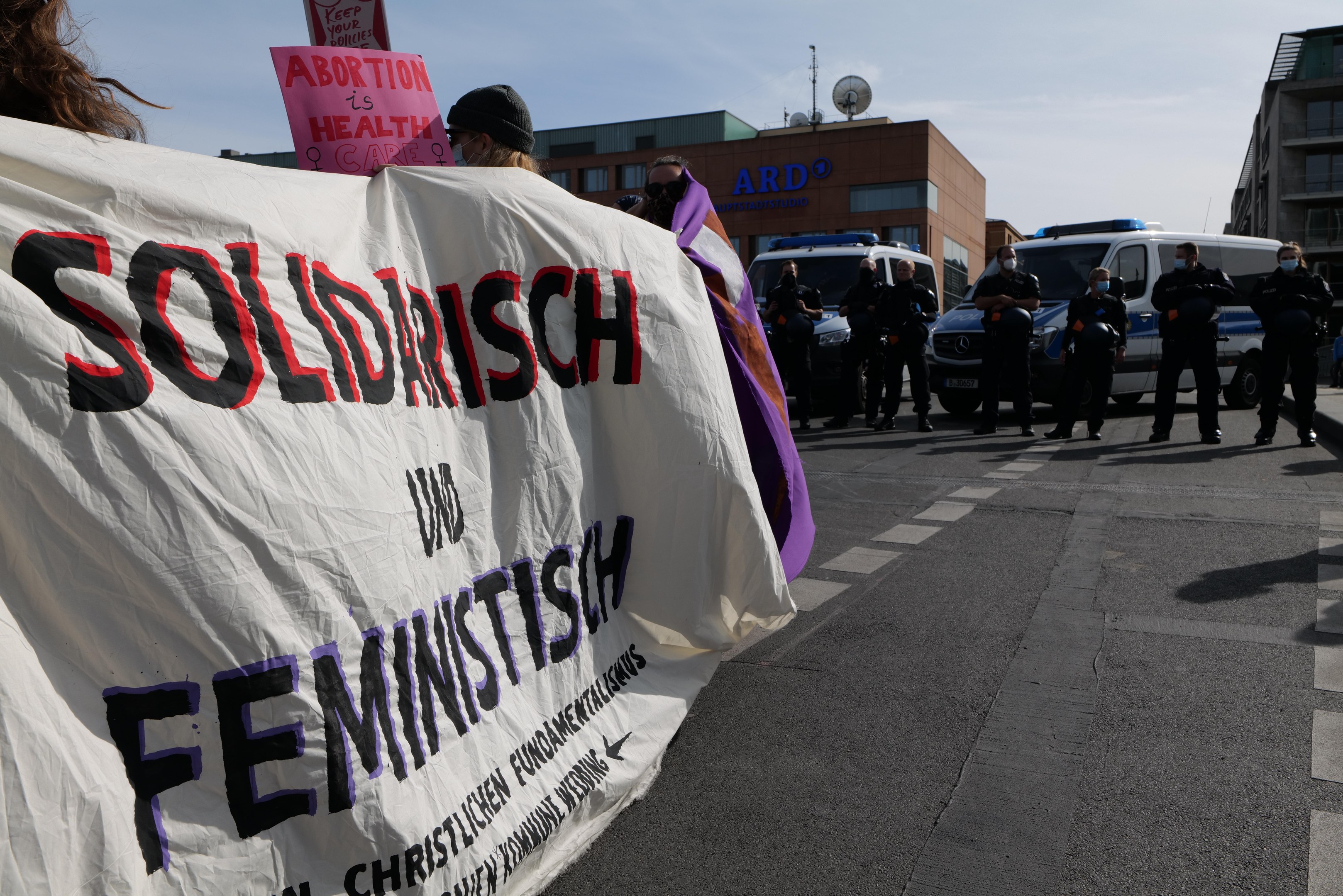 Eine Gruppe von Menschen marschiert auf einer Straße, hält ein 'Solidarität und Feminismus'-Schild, mit geparkten Fahrzeugen, Gebäuden, einer Schüsselantenne und einem klaren blauen Himmel im Hintergrund.