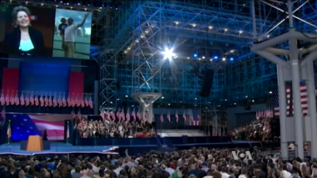 Eine große Menschenmenge sitzt vor einer Bühne mit einem Podium, Mikrofon, Fahnen, einem Bildschirm und Beleuchtung und wartet auf eine Präsidentschaftsdebatte.