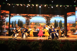 Eine Gruppe von Menschen in bunten Kostümen performs auf einer Bühne umgeben von Pflanzen, mit einem Bildschirm im Hintergrund und Lichtern, die von oben leuchten.
