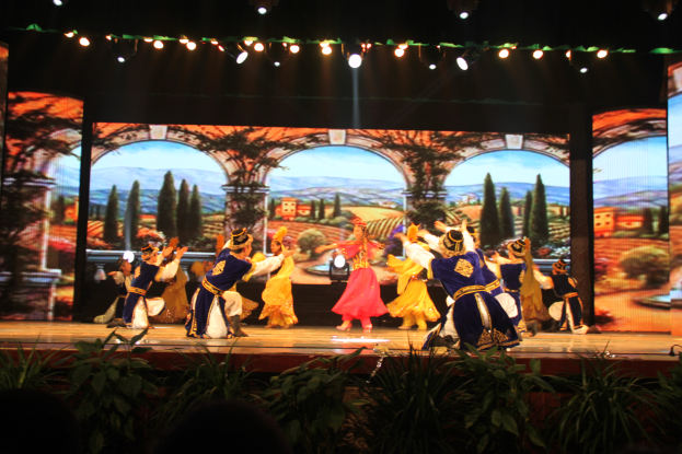 Eine Gruppe von Menschen in bunten Kostümen performs auf einer Bühne umgeben von Pflanzen, mit einem Bildschirm im Hintergrund und Lichtern, die von oben leuchten.