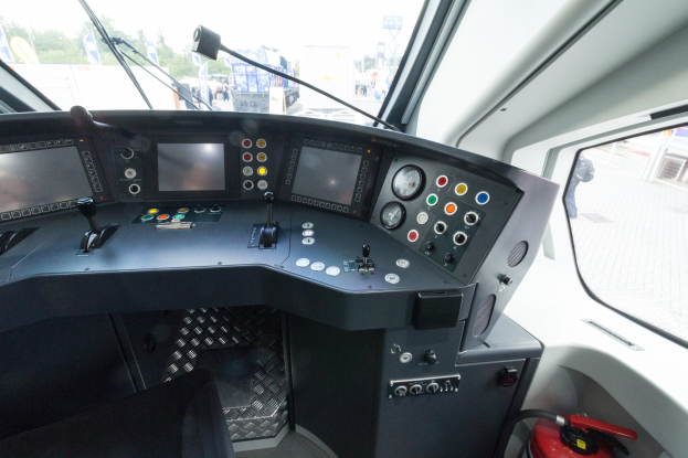 Cockpit einer Bahn mit einem Feuerlöscher im Vordergrund, das eine Glasfensteransicht von Menschen, Fahrzeugen, Bäumen, Mästäben und anderen Gegenständen außerhalb zeigt.
