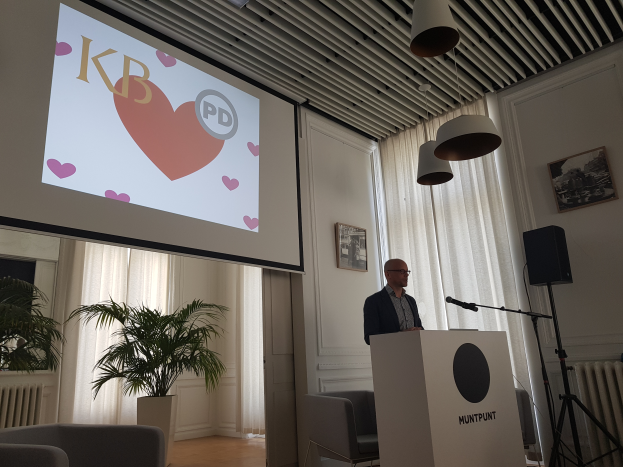 Ein Mann steht an einem Podium mit einem Mikrofon und hält eine Präsentation vor einem Projektionsschirm, mit Stühlen, einer Pflanze, Fotorahmen, Vorhängen und Deckenlampen im Hintergrund.