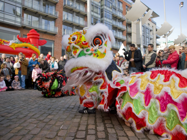 Ein lebendiges chinesisches Neujahrsfest in Amsterdam mit einem Löwen tanzen im Vordergrund mit einer Menge Menschen drumherum, einige halten Kameras, vor einem Hintergrund von Gebäuden, Laternenmasten und einem klaren blauen Himmel.