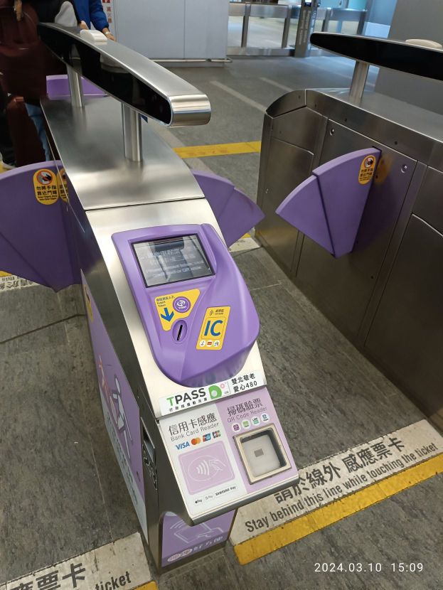 Eine Gruppe von Menschen vor einem Metalldetektor an einem Bahnhof, mit einem weiteren Metalldetektor rechts und einigen Menschen im Hintergrund.