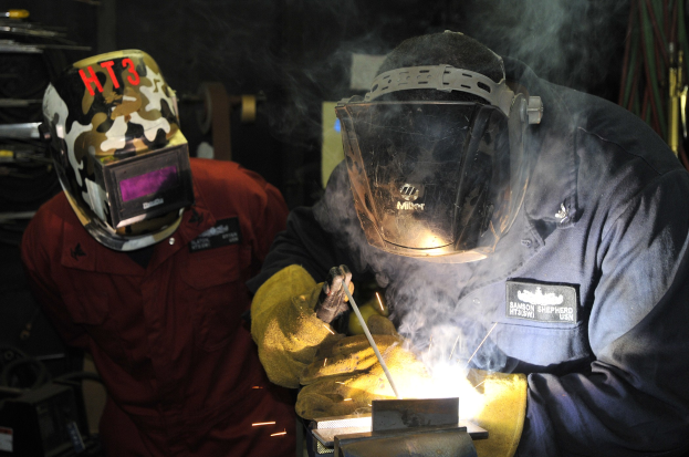 Zwei Schweißer in Helmen und Schutzausrüstung arbeiten an einem Metallstück, wobei Rauch aus dem Schweißprozess aufsteigt und die Werkstatt unordentlich ist.