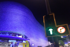 Das Selfridges Einkaufszentrum in Birmingham, England, ein großes Gebäude mit Säulen und Lichtern, umgeben von Bäumen, Laternenmasten und Fahrzeugen auf der Straße, mit einer Ampel im Vordergrund und dem Himmel im Hintergrund.