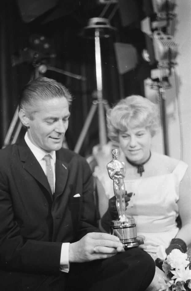 Ein Mann in einem schwarzen Blazer, einem weißen Hemd und einer Krawatte hölt eine Oscar-Statue, waehrend er neben einer Frau in formeller Kleidung sitzt, die einen Blumenstrau├č hölt, mit weicher Beleuchtung im Hintergrund.