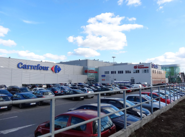 Großer Autohandel mit mehreren parkenden Fahrzeugen vorne, umgeben von städtischer Infrastruktur wie Gebäuden, Straßenlaternen, Schildern, fahrenden Verkehr und einem bewölkten Himmel im Hintergrund.