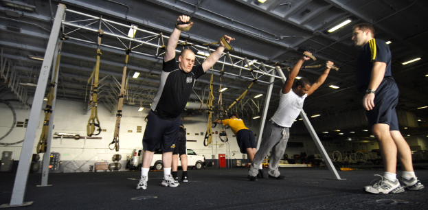 Gruppe von Männern, die an Metallstangen Klimmzüge machen, in einem Fitnessstudio mit Deckenbeleuchtung und verschiedenen Gegenständen im Hintergrund, ein Mann in der Mitte mit Schuhen.