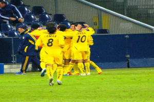 Eine Gruppe von Fußballspielern feiert ein Tor auf einem Rasenfeld, mit Zuschauern im Hintergrund auf Stühlen, Geländern und einer Wand.