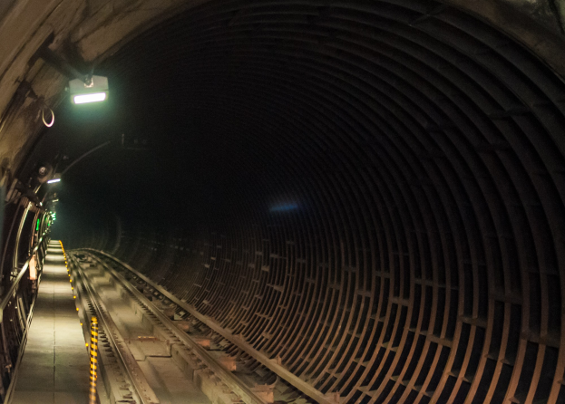 Ein Zug fährt durch einen schwach beleuchteten Tunnel in der Nacht, seine Bahn ist durch an der Decke montierte Lichter und sichtbare Schienen darunter markiert.