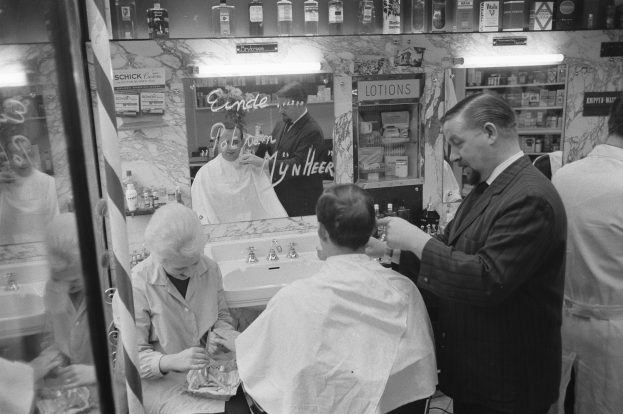 Ein Schwarz-Weiß-Foto eines Mannes, der steht und eine Schere hält, während eine Frau in einem Friseurstuhl sitzt und ein Tuch um die Schultern trägt, mit einem Waschbecken, Spiegel, Regalen mit Flaschen und einer Glastür im Hintergrund.