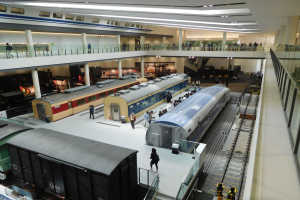 Eine Museumsausstellung zeigt verschiedene Züge in unterschiedlichen Größen und Farben, die von Menschen auf dem Boden, Geländern und Säulen unter Deckenlampen betrachtet werden.