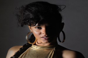 Eine Frau in einem goldenen Kleid und großen Ohrringen steht selbstbewusst vor einer Wand, ihre glatten Bob-Haare sind ordentlich frisiert, sie trägt dezentes Make-up mit pinkem Rouge und glänzenden Lippen und schaut direkt in die Kamera mit einem entschlossenen Gesichtsausdruck.