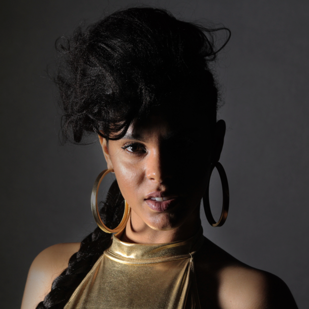 Eine Frau in einem goldenen Kleid und großen Ohrringen steht selbstbewusst vor einer Wand, ihre glatten Bob-Haare sind ordentlich frisiert, sie trägt dezentes Make-up mit pinkem Rouge und glänzenden Lippen und schaut direkt in die Kamera mit einem entschlossenen Gesichtsausdruck.