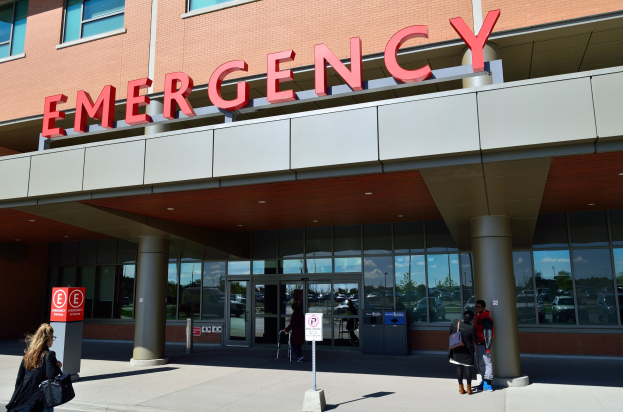 Gruppe von Menschen, die vor einem Notaufnahmesgebäude mit Glaswänden, Säulen, einer Namensplakette, Hinweistafeln und einem Mülleimer gehen.