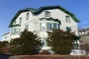 Ein weißes Haus mit grüner Umrandung und Dach, umgeben von Bäumen, Pflanzen und einem Zaun, mit einer Straße im Vordergrund und einem klaren blauen Himmel im Hintergrund.