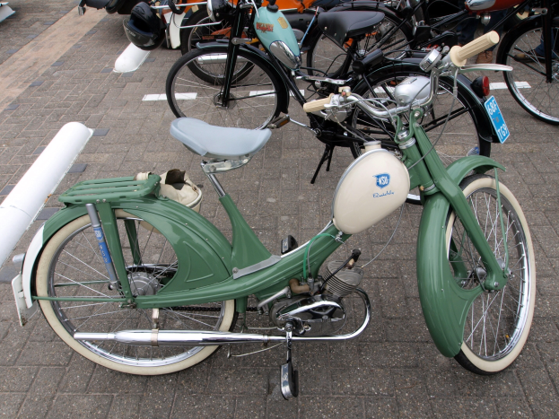 Ein grünes Moped mit einem weißen Rahmen und schwarzem Sitz neben anderen Motorrädern auf einem Gehweg geparkt, mit ein paar Menschen in der Nähe.