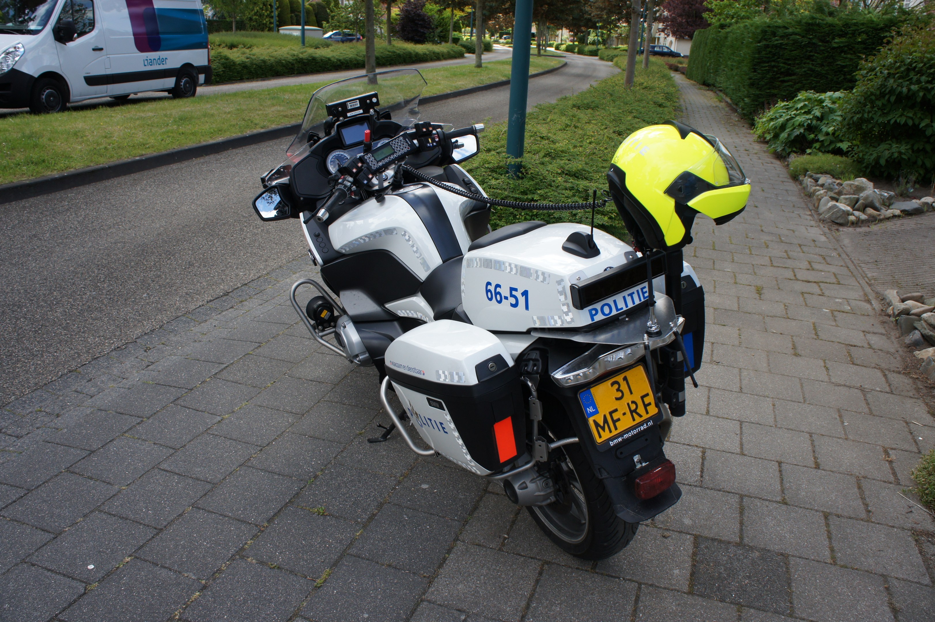 Polizeimotorrad am Straßenrand mit Helm darauf, umgeben von Bäumen, Polen, Gebäuden, Pflanzen, Gras, Steinen und anderen Fahrzeugen im Hintergrund.