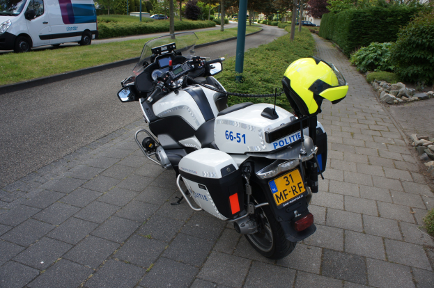 Polizeimotorrad am Straßenrand mit Helm darauf, umgeben von Bäumen, Polen, Gebäuden, Pflanzen, Gras, Steinen und anderen Fahrzeugen im Hintergrund.
