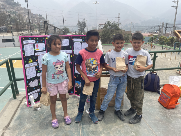 Gruppe von Kindern auf einem Basketballfeld mit Papiertüten in den Händen, mit Gegenständen auf der rechten Seite und einer Tafel mit Text auf der linken Seite, vor einer Außenkulisse mit Zaun, Pfosten und Gebäuden.
