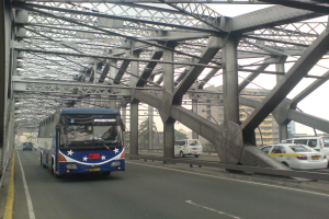 Ein blauer Bus fährt über eine Brücke mit Autos, umgeben von Gebäuden, Bäumen, Pfählen und einem klaren blauen Himmel.