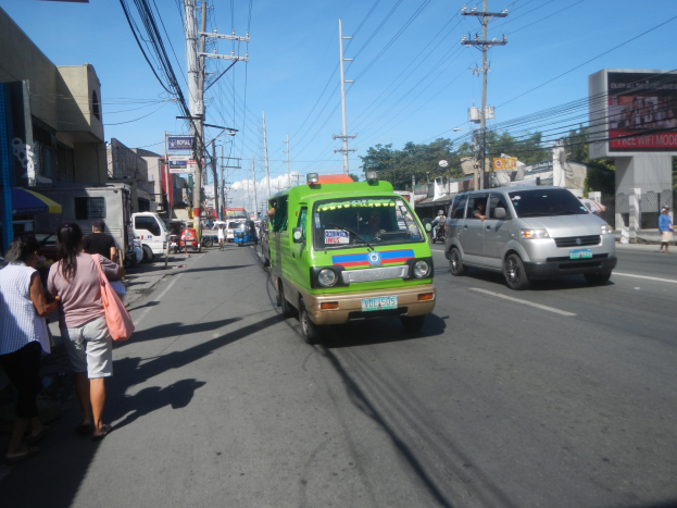 Ein grüner Lieferwagen fährt eine von hohen Gebäuden gesäumte Straße entlang, mit Passanten auf dem Gehweg, Oberleitungen, Bäumen in der Ferne und einem klaren blauen Himmel.