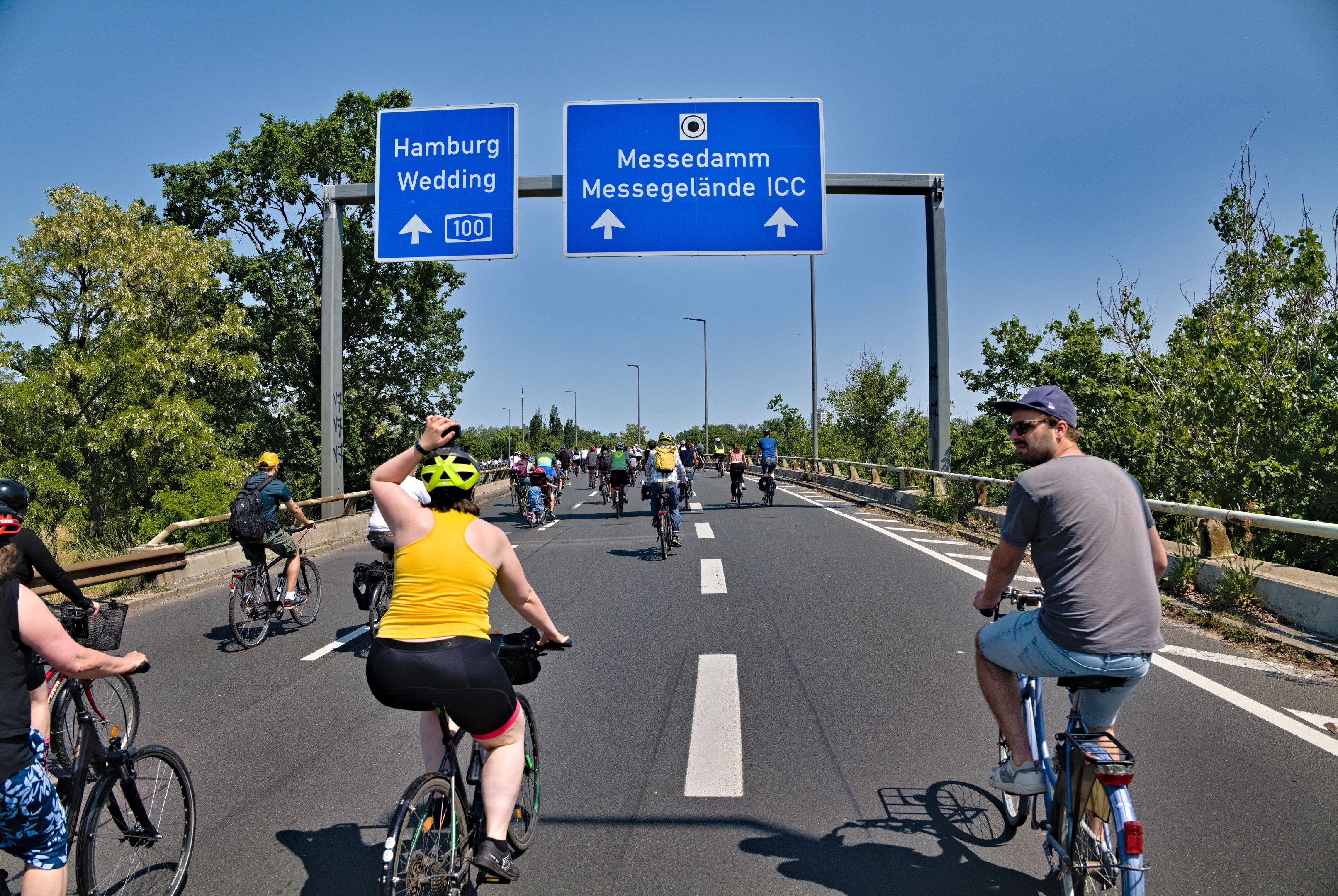 Eine Gruppe von Menschen in Helmen, die Fahrräder auf einer Straße mit Bäumen auf einer Seite und Laternen im Hintergrund unter einem klaren blauen Himmel fahren, mit einer Tafel, die eine Fahrradtour in Hamburg anzeigt.