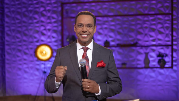 Stephen Colbert, der in Anzug, Krawatte und Brille auf einer Bühne steht und ein Mikrofon hält, mit einer Wand, einem Ständer, einer Lampe und verschiedenen Gegenständen dahinter.